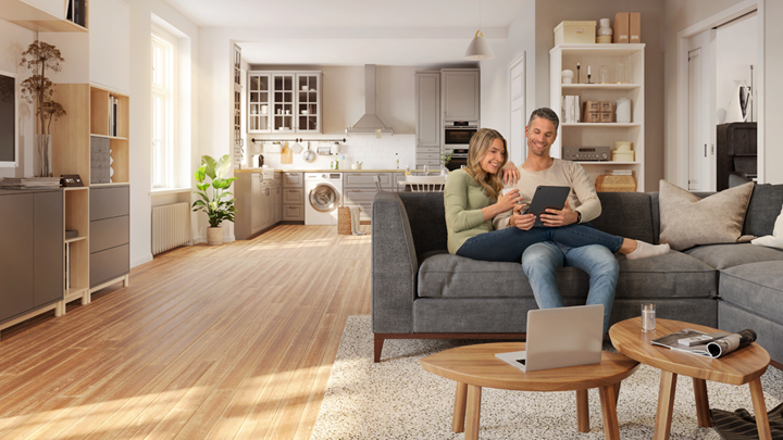 Junges Paar sitzt auf Sofa und schaut in ein Tablet, im Hintergrund Küche und Wohnzimmerschrank mit Hausrat